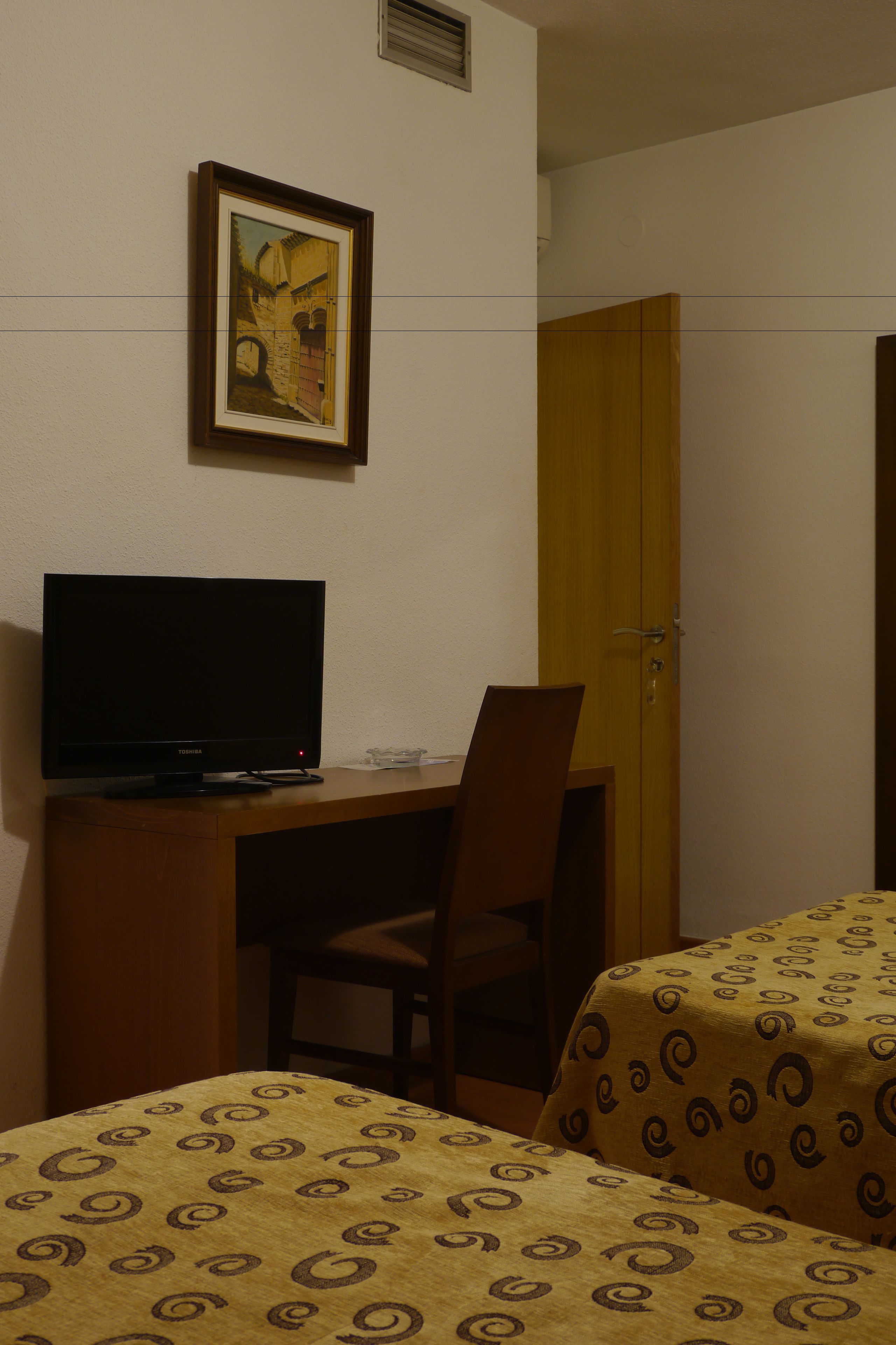 A hotel room with a desk, a television, a bed with patterned bedding, a door, and a framed picture on the wall.