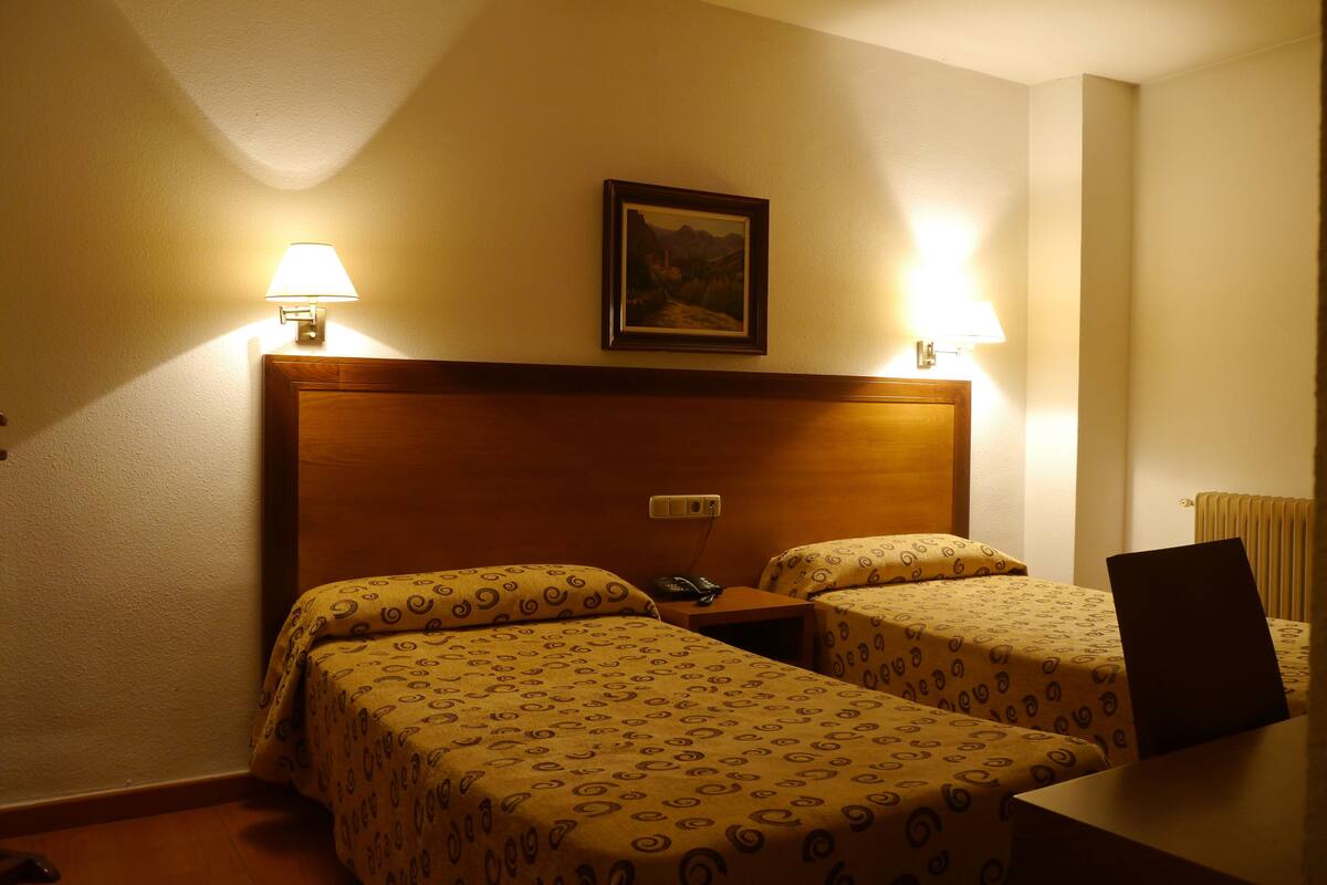 Habitación de hotel con dos camas individuales, un cabecero de madera, lámparas de noche, un escritorio con una computadora portátil y un cuadro en la pared.