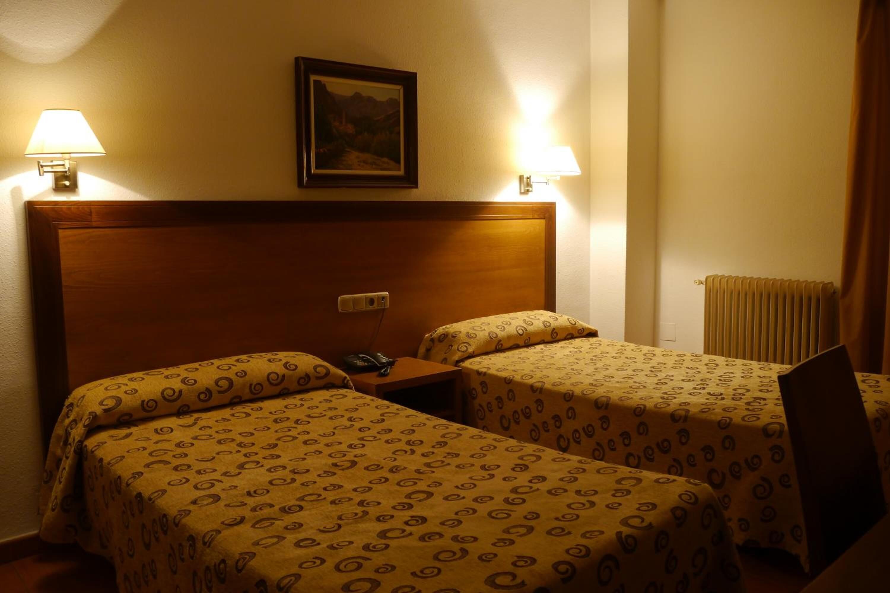 A hotel room with two beds, a wooden headboard, a nightstand with a phone, and two wall-mounted lamps.