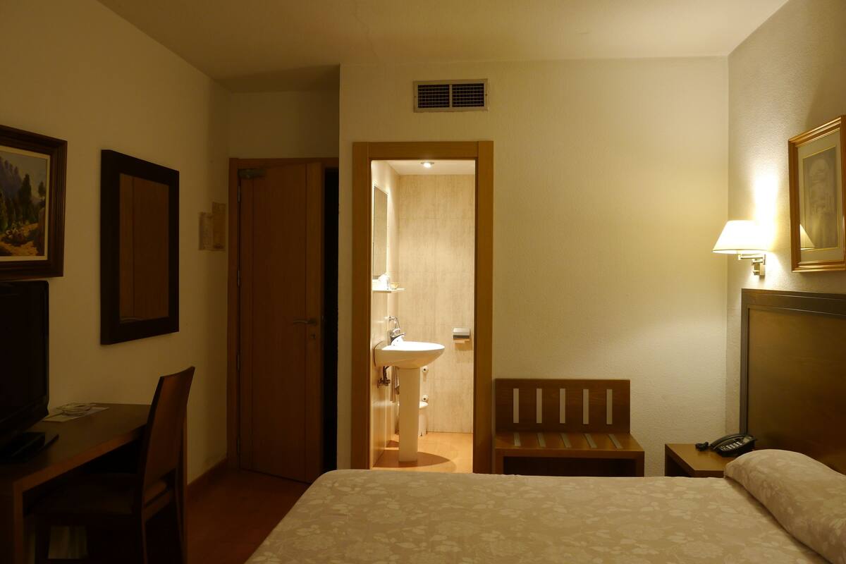 Habitación de hotel con cama, escritorio, silla, mesita de noche con lámpara, teléfono y baño visible a través de una puerta abierta.