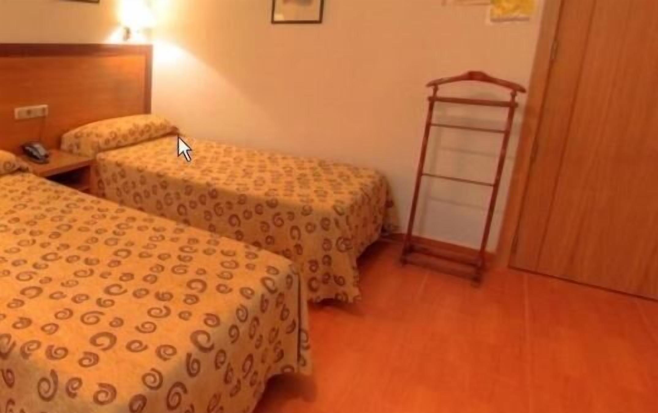 A room with two beds, a wooden wardrobe, and a small table with a lamp.