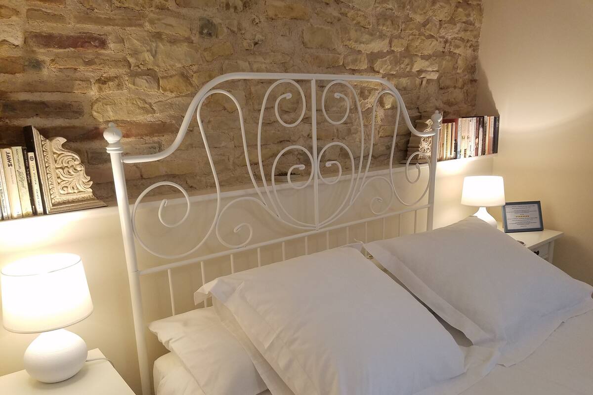 Una cama bien hecha con sábanas y almohadas blancas, un cabecero de metal blanco, una mesita de noche con lámpara y un plato decorativo, y una pared con textura de piedra y una estantería llena de libros.