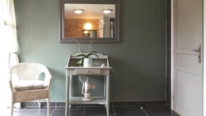 Una habitación con una silla de mimbre blanca, una consola blanca con una planta y un objeto decorativo, un espejo en la pared y una puerta a la derecha.