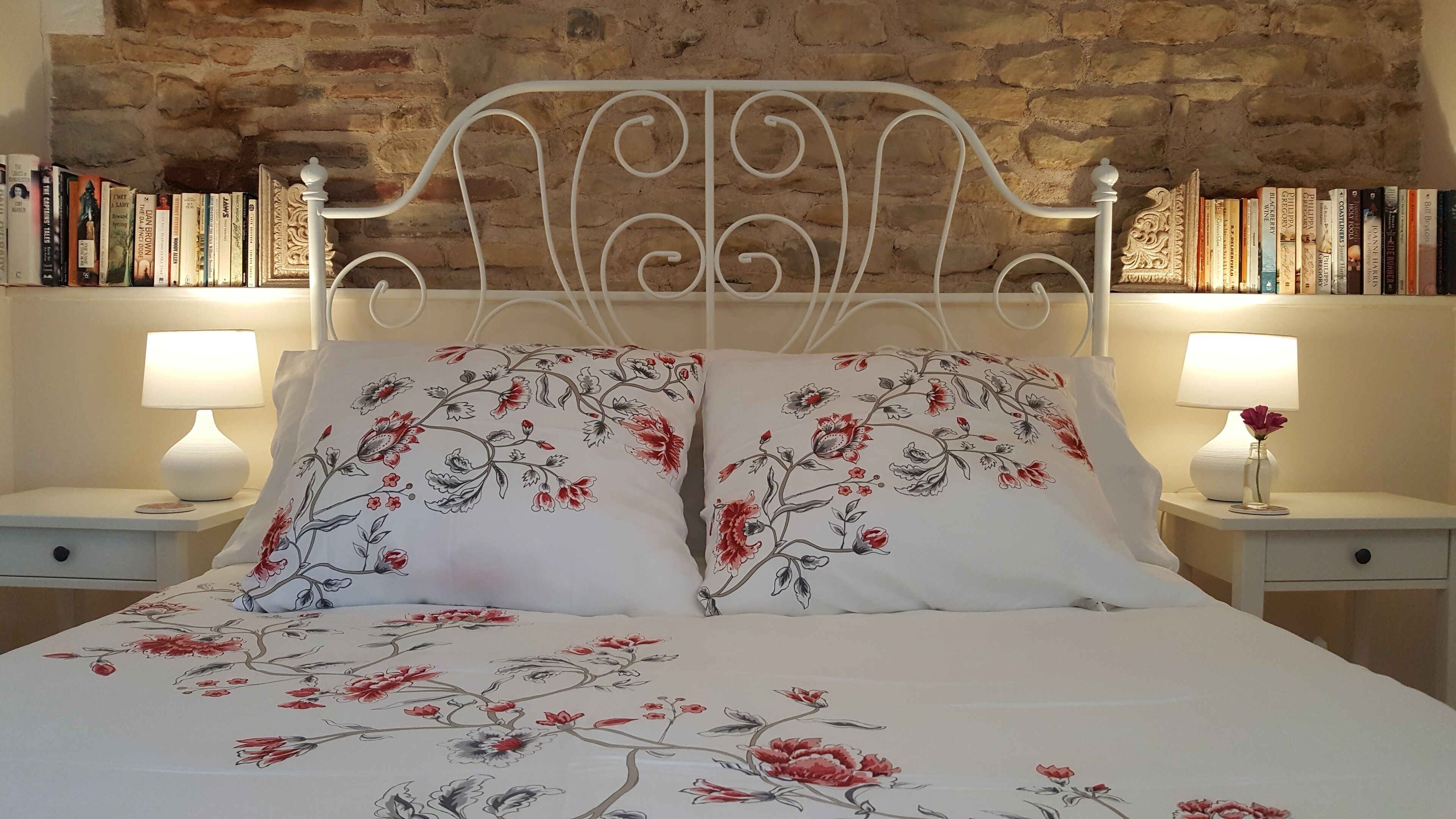 Una cama con ropa de cama floral, dos lámparas de noche y una estantería con libros detrás de un muro de piedra.