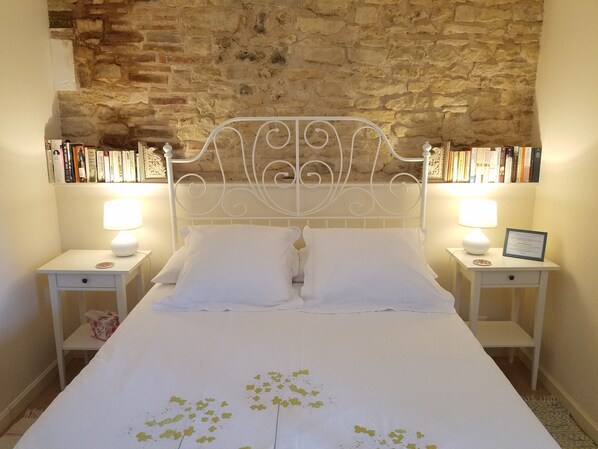 Una cama bien tendida con sábanas blancas y motivos de hojas verdes decorativas, flanqueada por dos mesitas de noche con lámparas y certificados enmarcados. Se aprecia una pared de piedra y una estantería con libros al fondo.