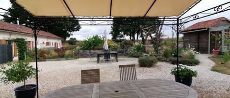 A covered patio area with a table and chairs, surrounded by gravel and potted plants.
