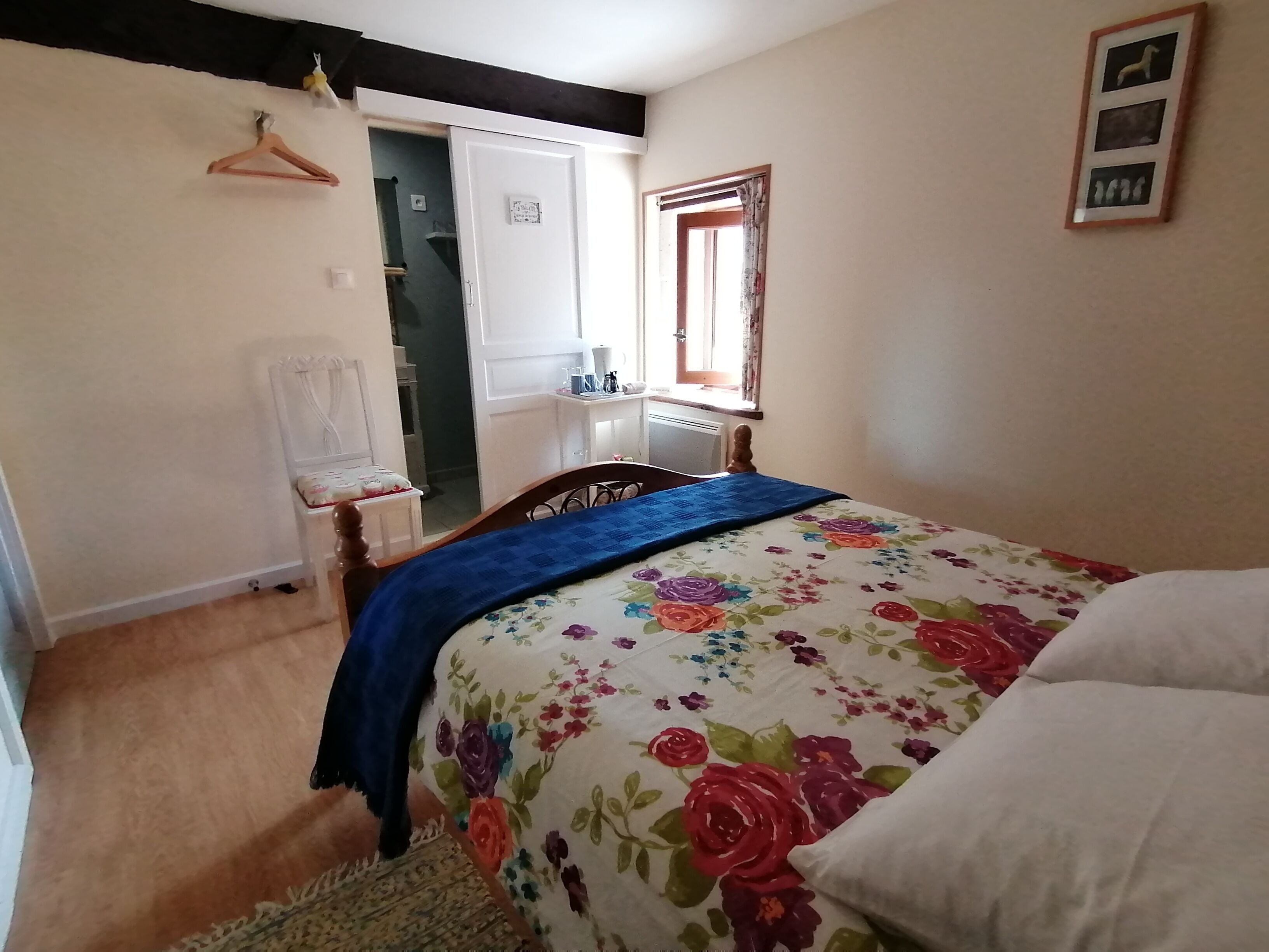 Quarto com colcha florida, uma cadeira de madeira, escrivaninha com abajur e janela com cortinas.