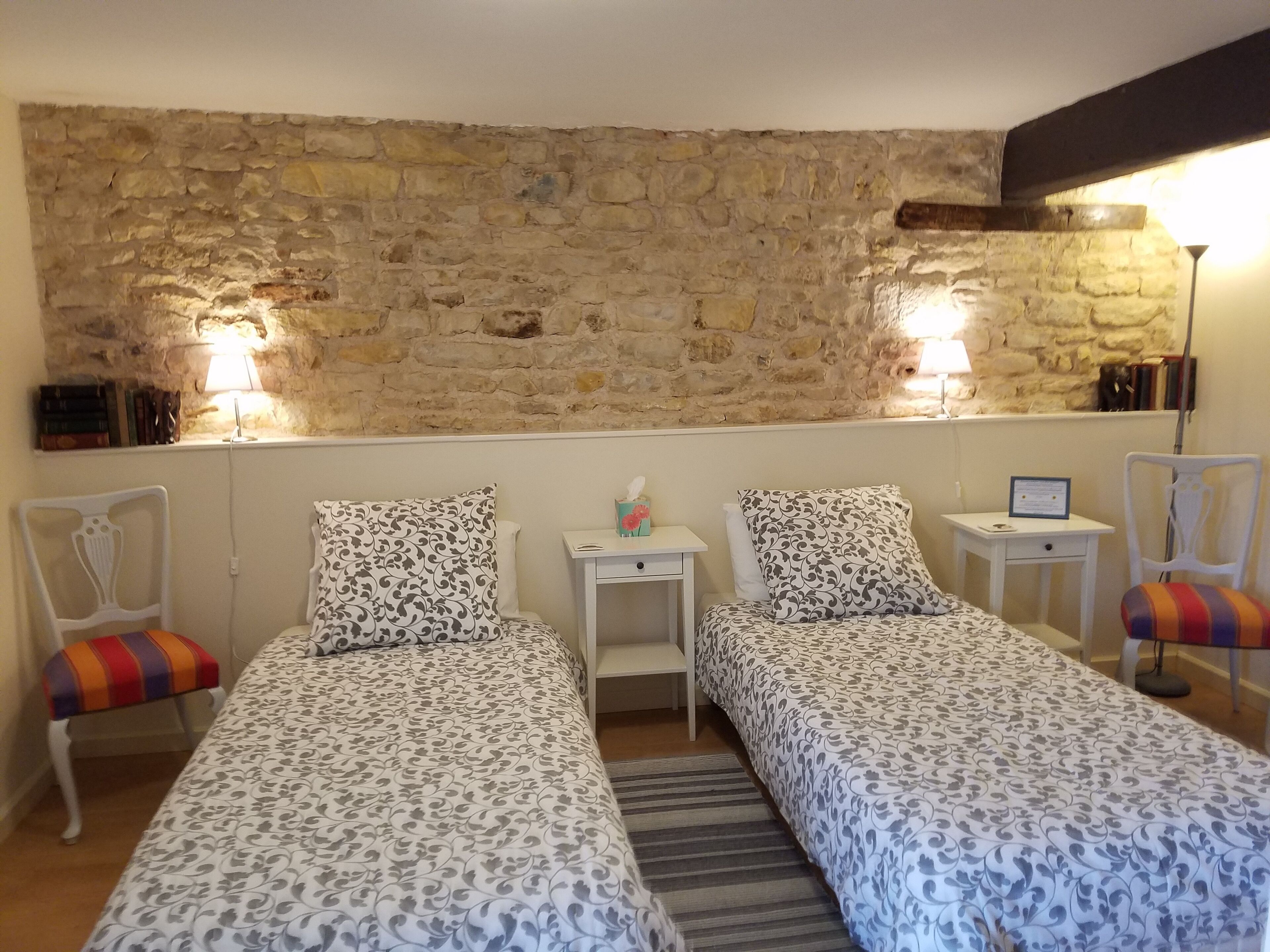 A room with two single beds, a small table, a chair, a lamp, and a stone wall.