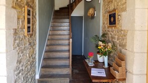 Una escalera de madera conduce a una habitación con pared de piedra, un reloj y una pequeña mesa con un cuaderno y un jarrón de flores.