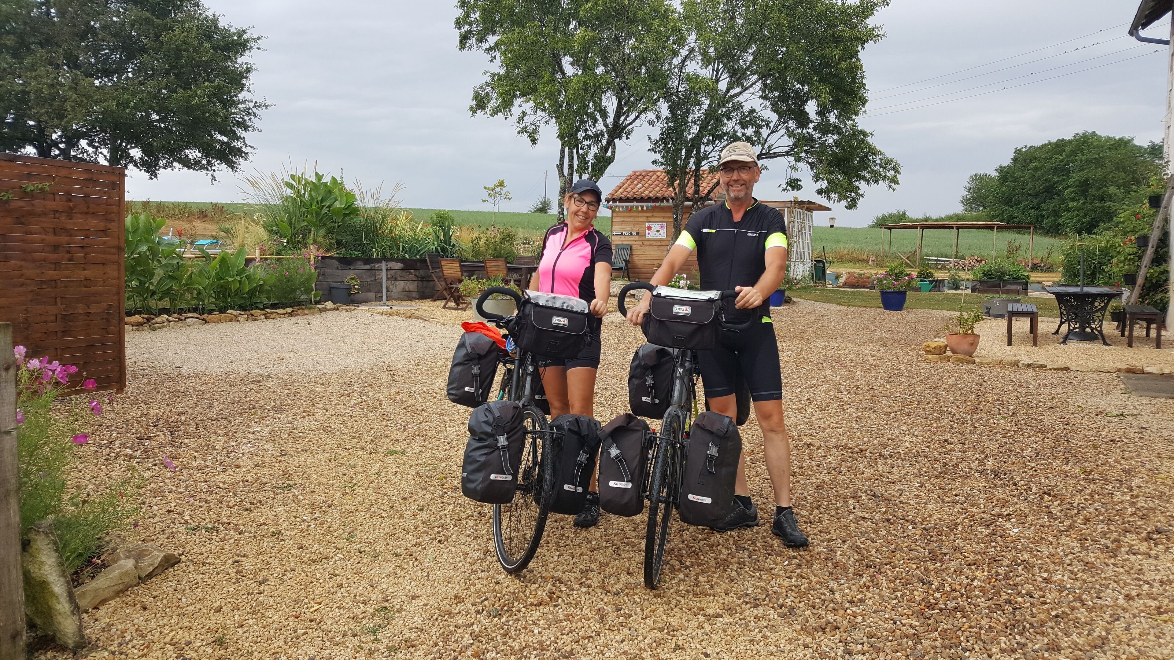 Two people standing next to bicycles with bags attached, in a gravel area with trees and a wooden fence in the background.
