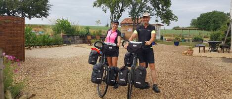 Two people standing next to bicycles with bags attached, in a gravel area with trees and a wooden fence in the background.