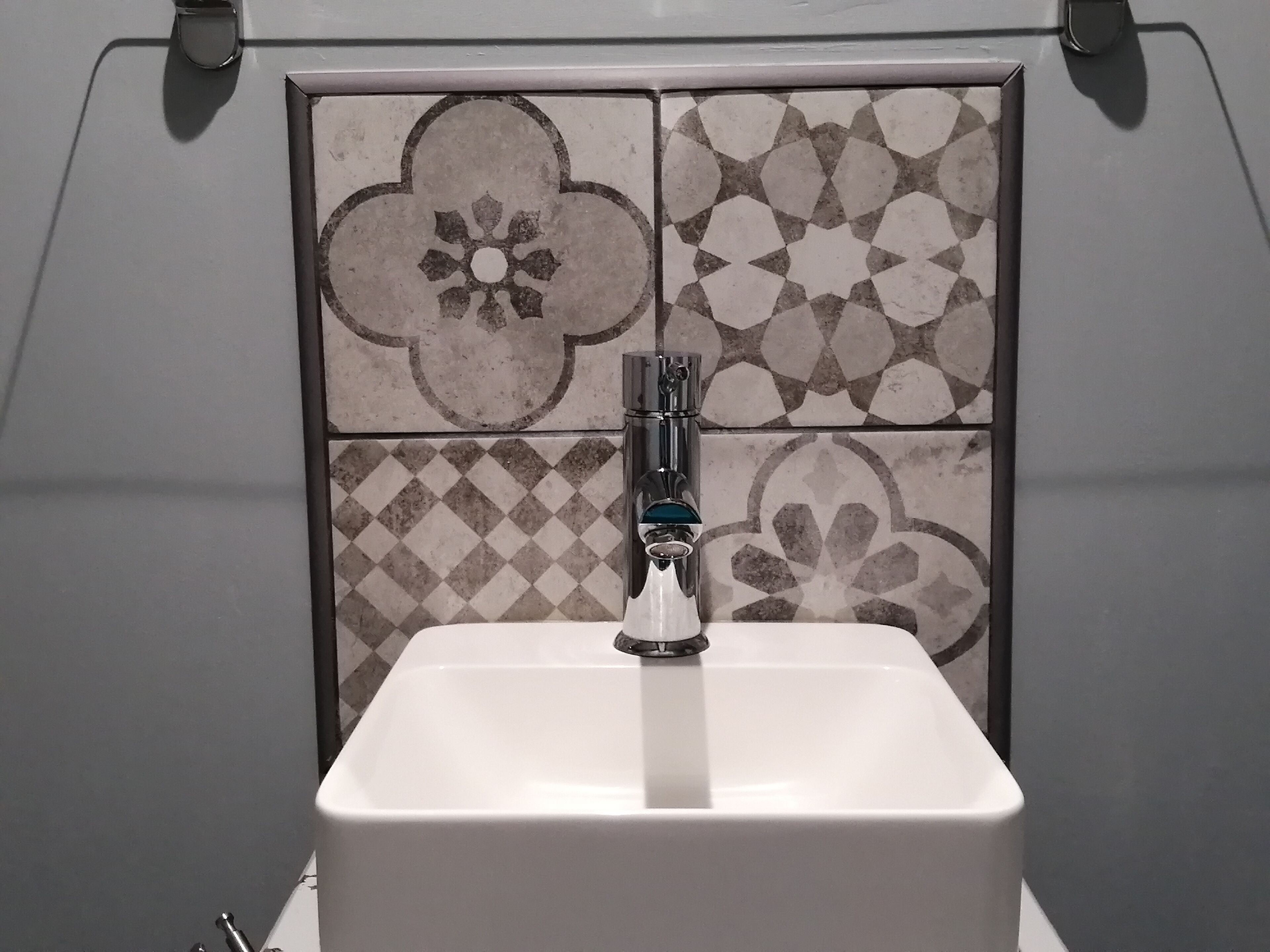 Un bagno con un lavandino bianco, un rubinetto moderno e una parete piastrellata con motivi geometrici e floreali.