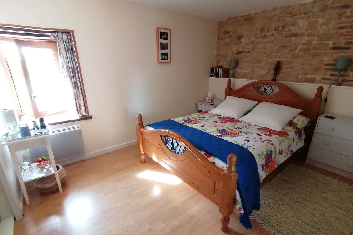 Un dormitorio con una cama de madera, una mesita de noche, un escritorio, una ventana con cortinas y una pared de piedra.