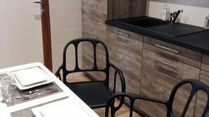 A compact kitchen with a built-in oven, a black countertop, and wooden cabinets. There are two black chairs and a dining table set with plates, glasses, and cutlery. - What Else Hotel (Saint-Vulbas)