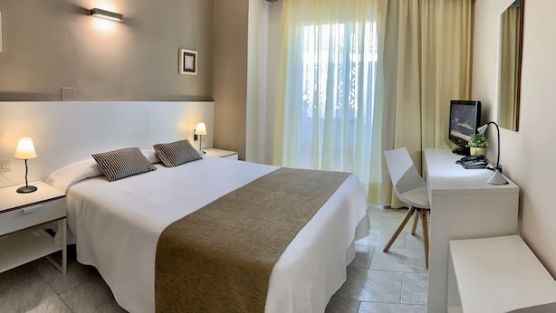Une chambre d’hôtel avec un grand lit, des tables de chevet avec des lampes, un bureau avec une chaise, une télévision et une fenêtre avec des rideaux.