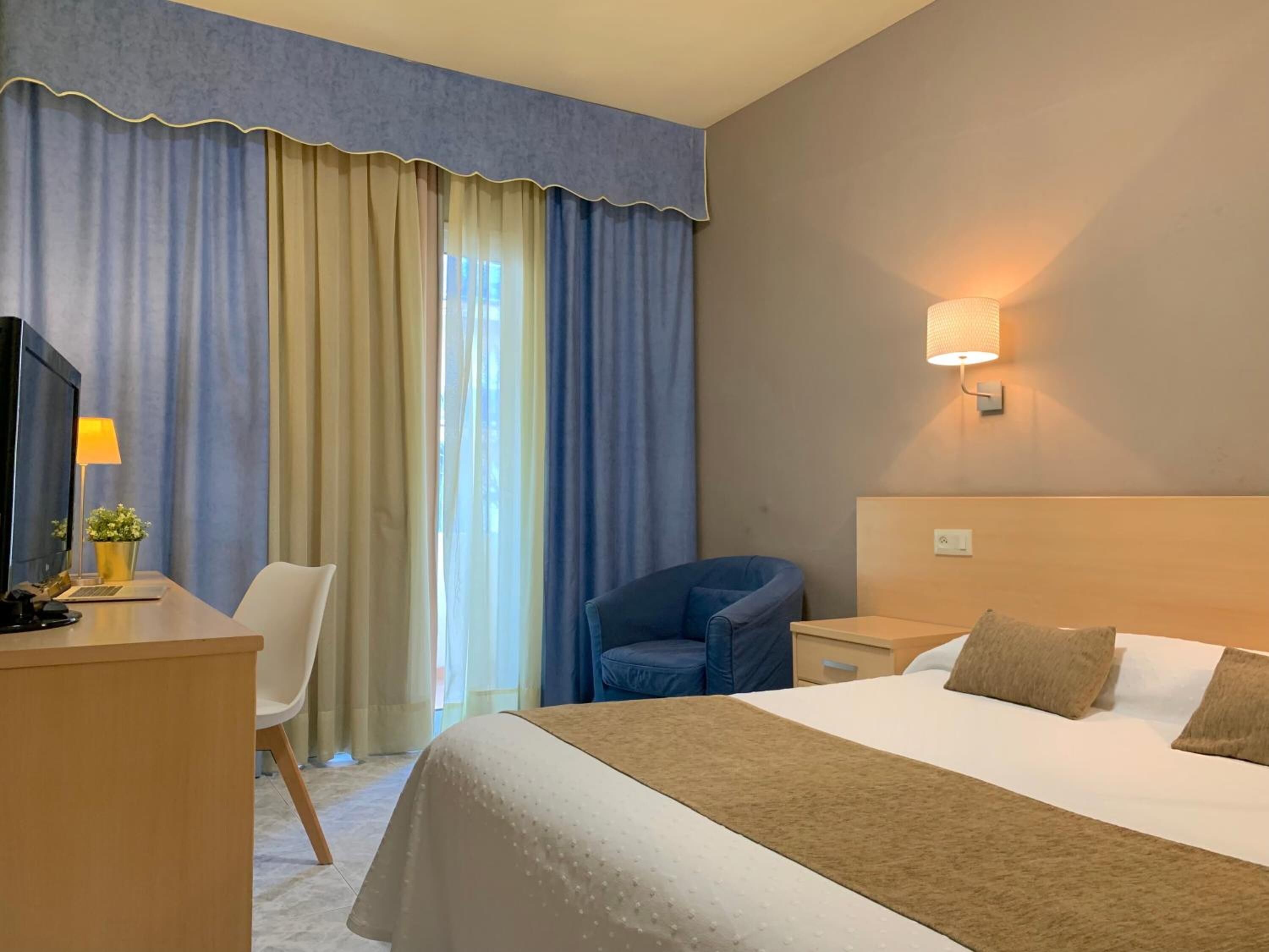 A hotel room with a bed, a desk with a computer, a chair, a lamp, and a window with curtains.