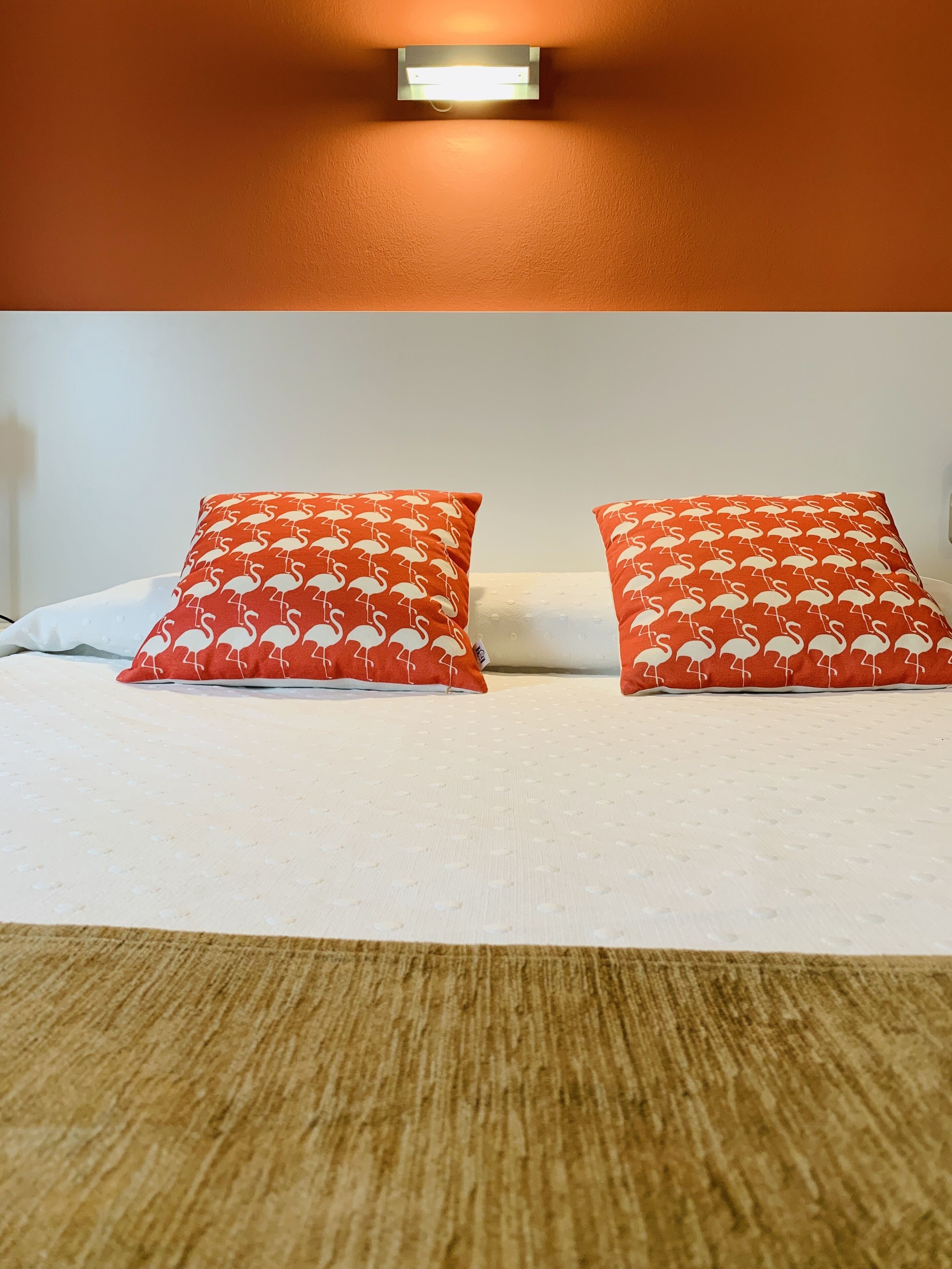 A hotel room with a bed, two red pillows with a flamingo pattern, a wall-mounted light, and a textured brown carpet.