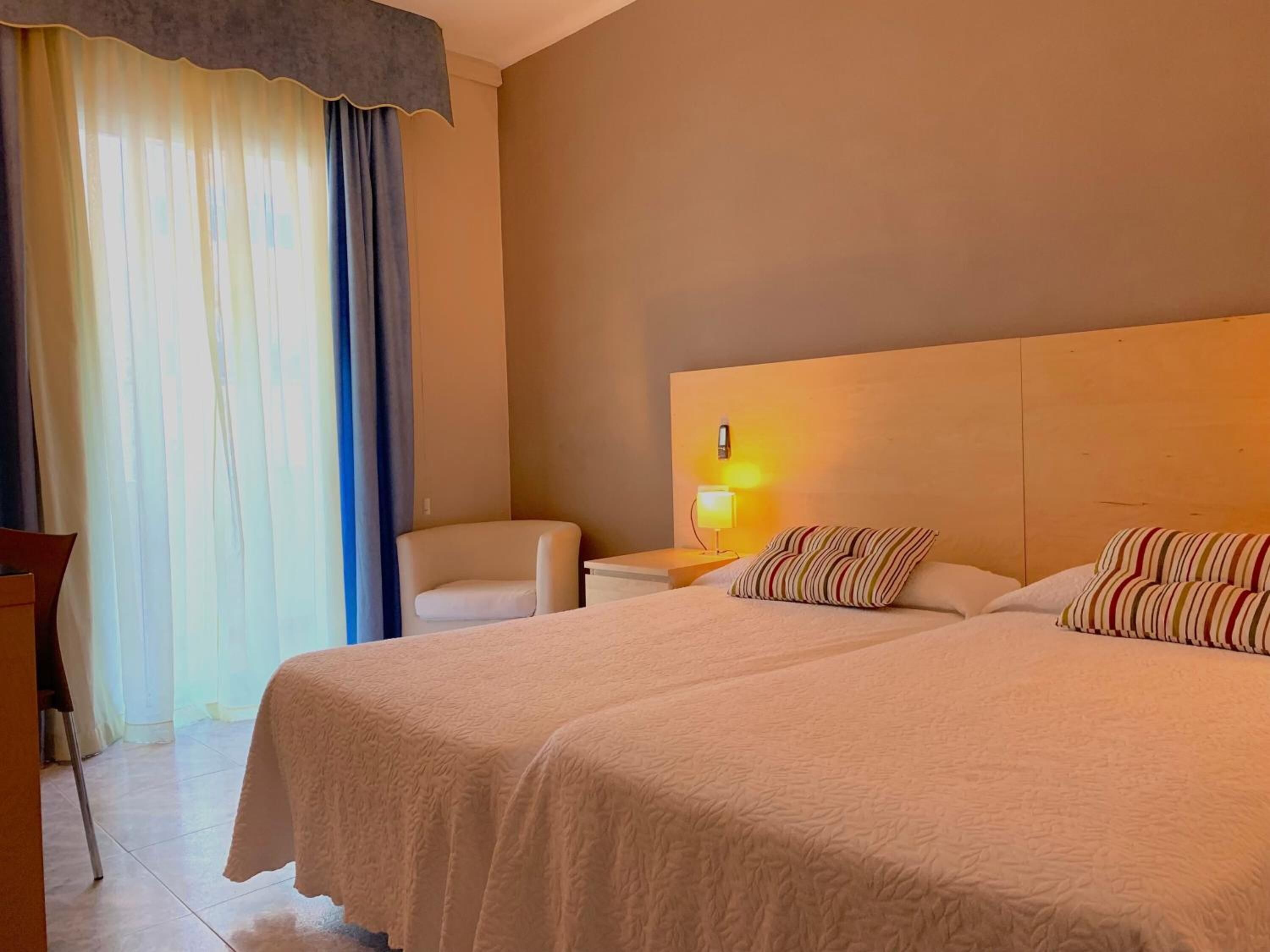 Een hotelkamer met twee bedden, een stoel, een nachtkastje met een lamp en een raam met gordijnen.