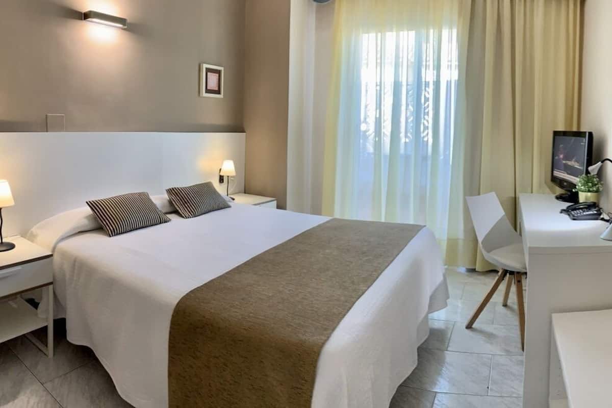 A hotel room with a bed, bedside tables, a desk, a chair, a TV, and a window with curtains.