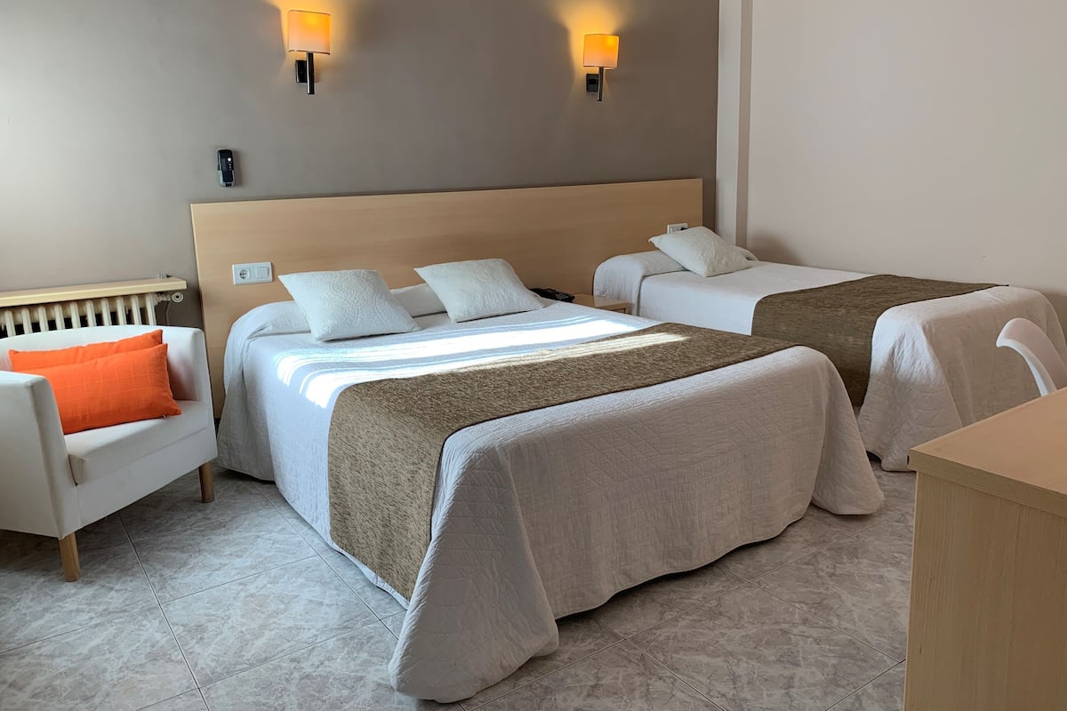 A hotel room with two beds, a chair, a bedside table, and two wall-mounted lamps.