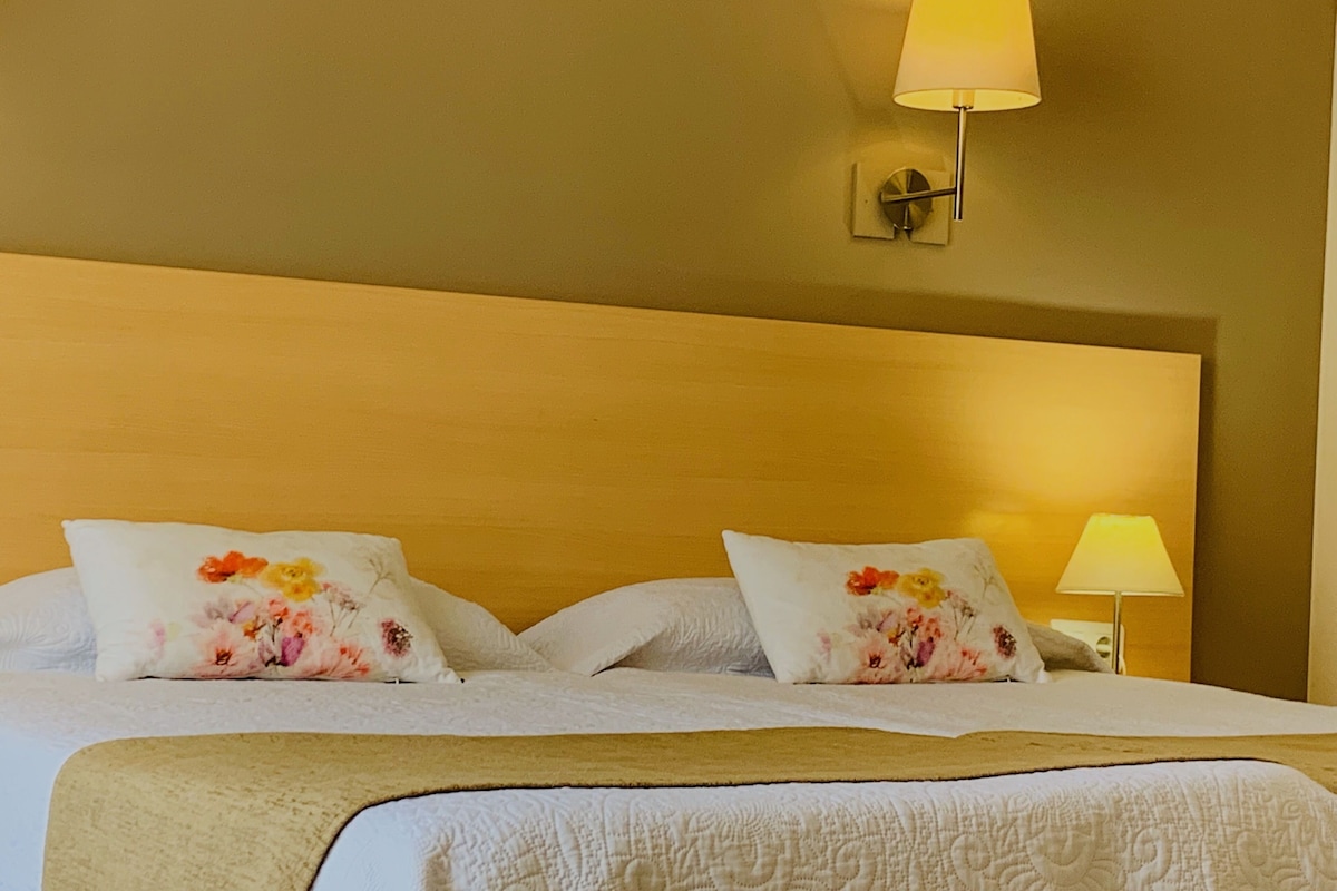 Une chambre d’hôtel avec un grand lit, deux oreillers ornés de motifs floraux, une lampe de chevet et un luminaire fixé au mur.