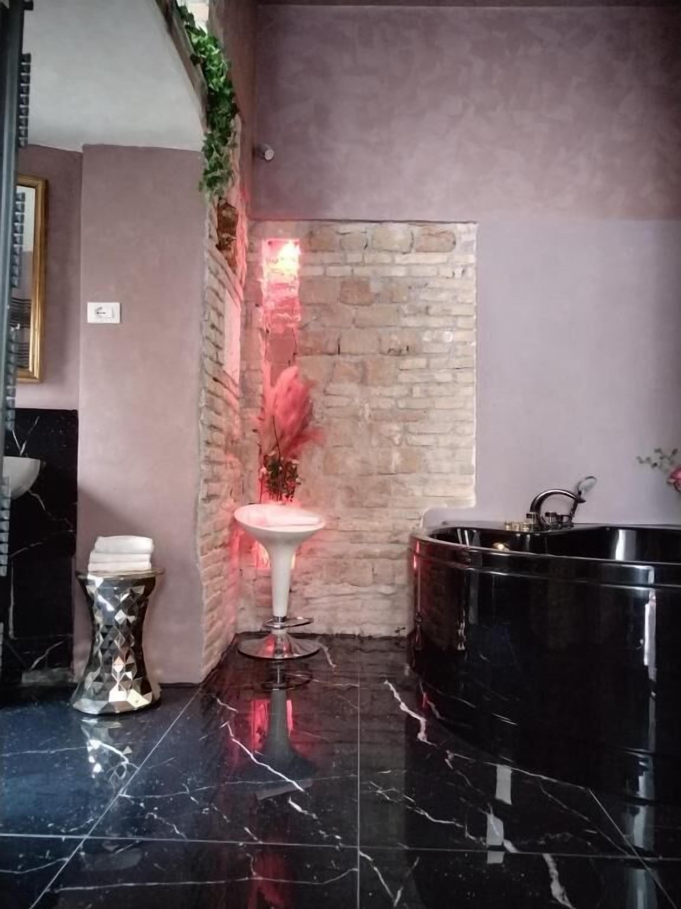 Un baño con piso de mármol negro, un lavamanos blanco pedestal, una bañera negra grande y una pared de acento de ladrillo.
