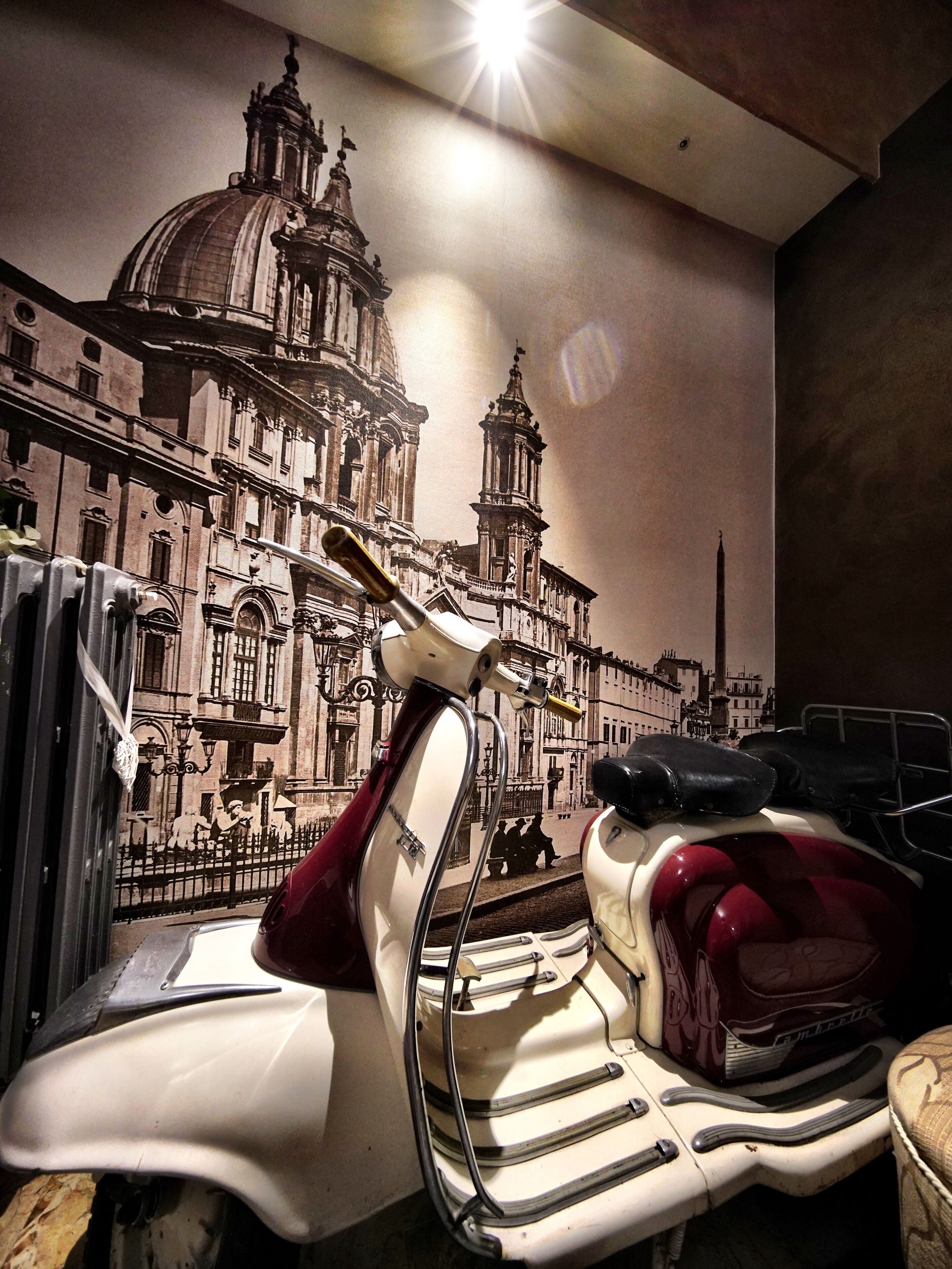 A room with a wall mural of a historic building, a white and red scooter, and a black chair.
