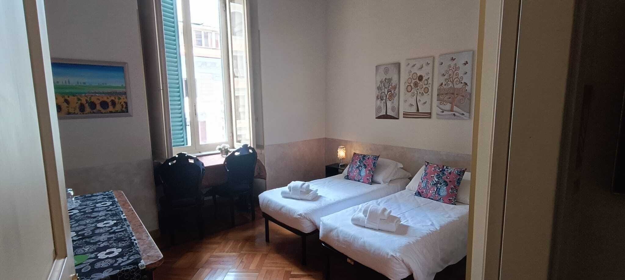 Egy szállodai szoba két ággyal, egy kis asztallal, székekkel és egy ablakkal, melynek redőnyei vannak.