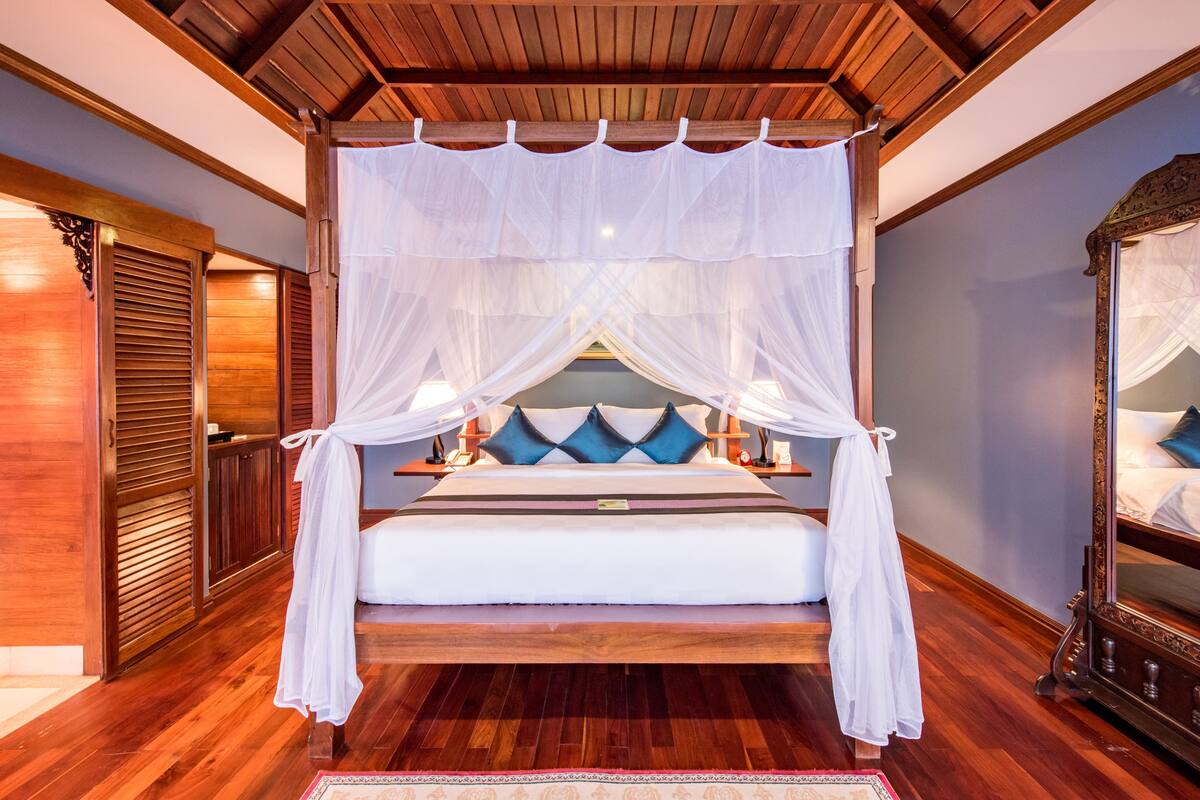 A bedroom with a wooden floor, a canopy bed, and a nightstand with a lamp.