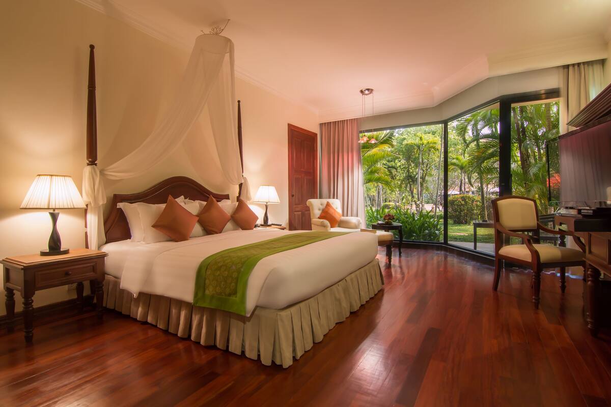 A spacious bedroom with a large bed, a sitting area with a chair, and a view of greenery through large windows.