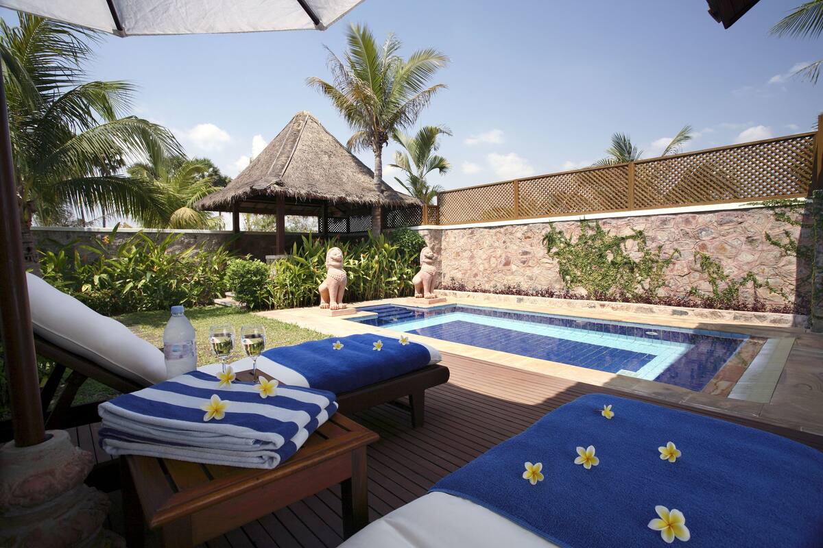 A poolside area with lounge chairs, a thatched-roof structure, and a stone wall with greenery.