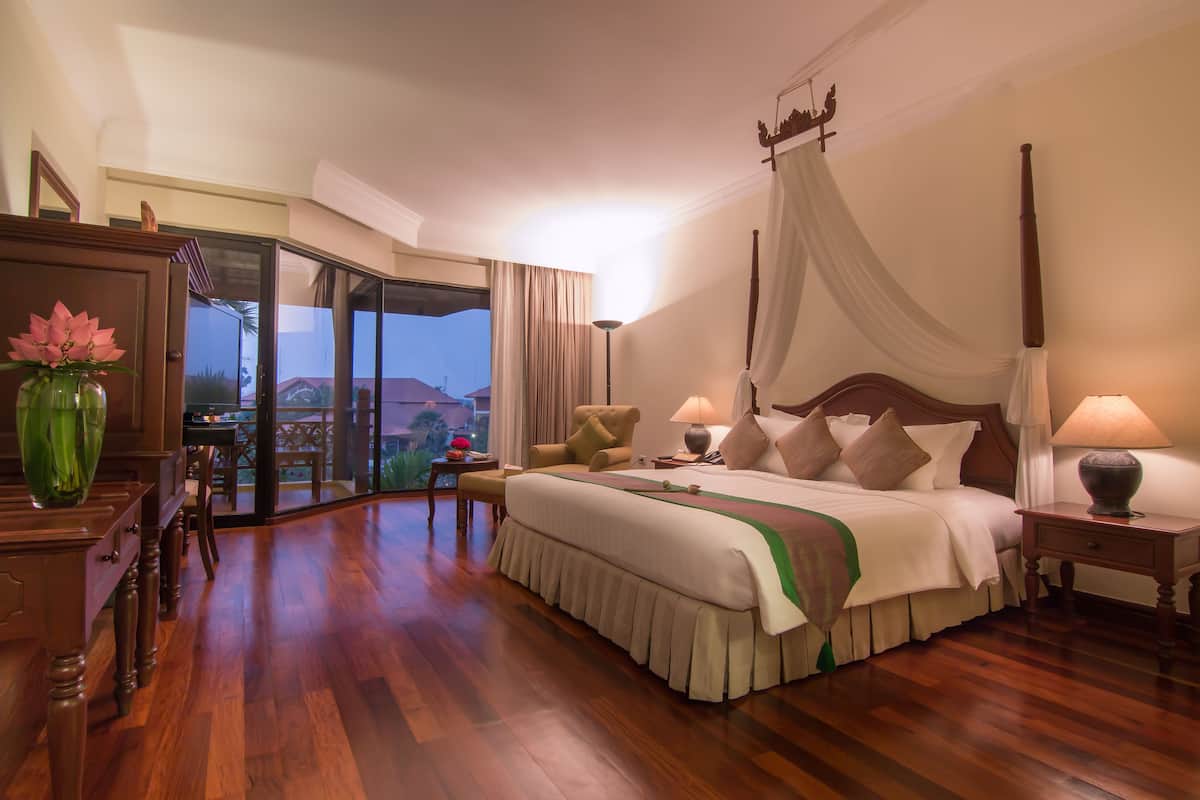 A spacious bedroom with a large bed, a sitting area with a chair, a wooden dresser, and a view of the outdoors.