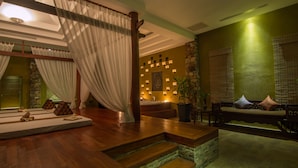 A room with a wooden floor, a bed with white curtains, a stone wall with lights, a bench, and a sofa with cushions. - Angkor Palace Suite & Villa (Siem Reap)
