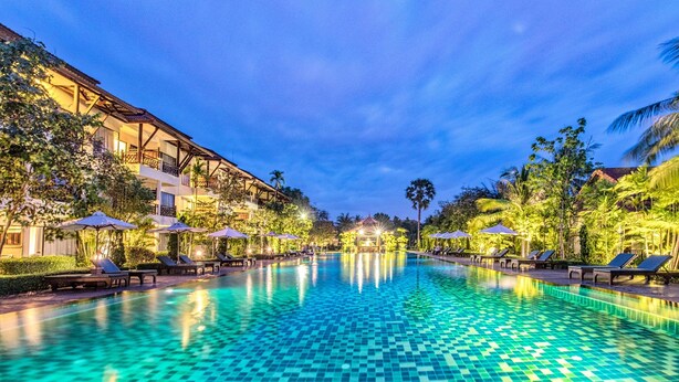 A large, clear swimming pool with lounge chairs and umbrellas, surrounded by lush greenery and a building with balconies. - Angkor Palace Suite & Villa (Siem Reap)
