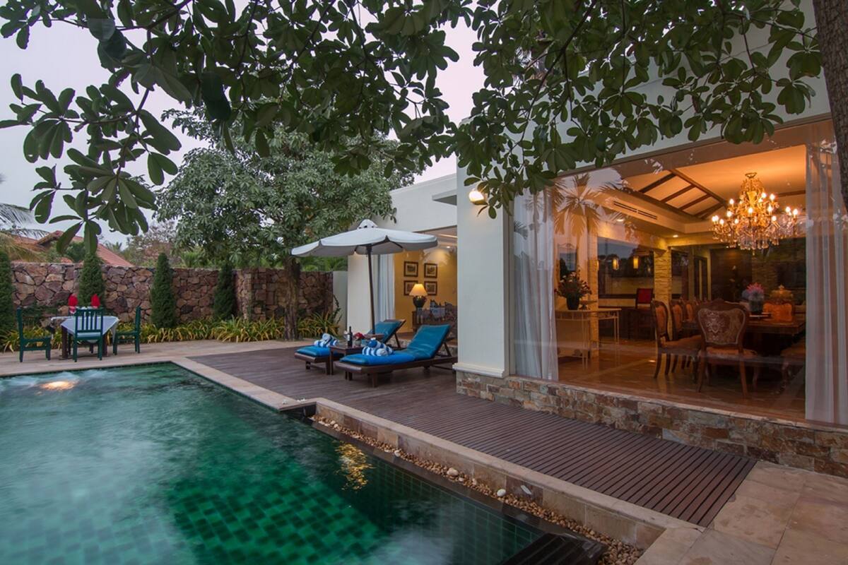 A pool area with lounge chairs, a dining area with chairs and tables, and a view of the interior through glass doors.
