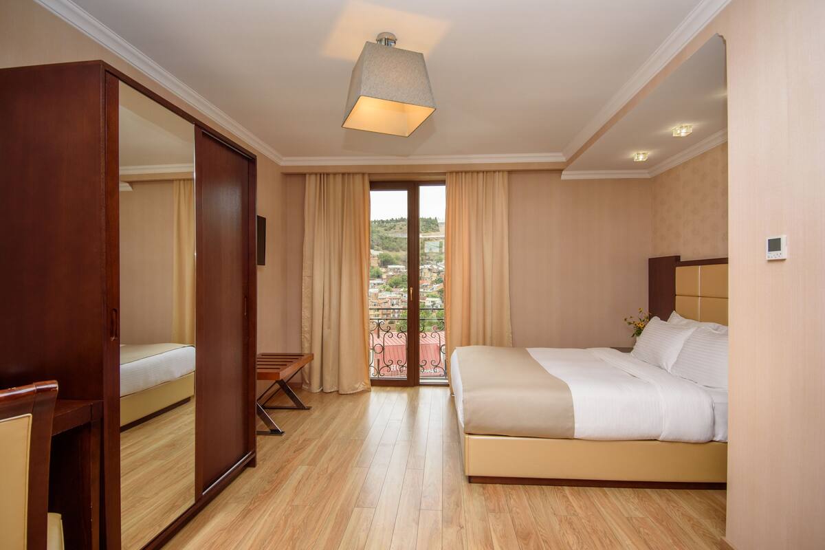 A hotel room with a bed, a wooden wardrobe, a desk, and a balcony with a view of the city.