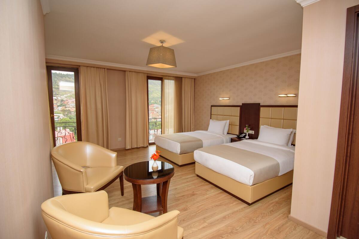 A hotel room with two beds, a sitting area with chairs, a small table with a vase, and a view of the outside.