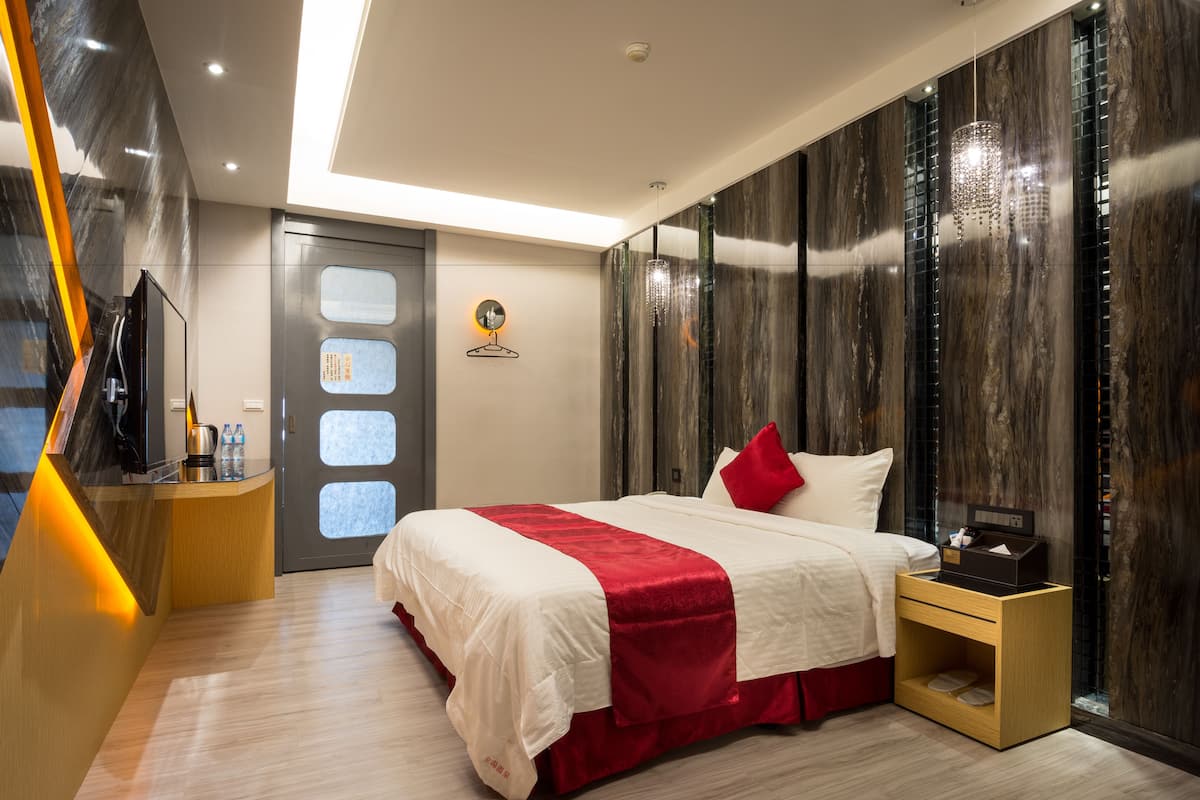A modern hotel room with a large bed, a wooden nightstand, a desk with a computer, a TV, and a wall-mounted clock.
