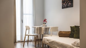 A small, well-lit room with a bed, a round dining table with chairs, a potted plant, and a painting on the wall.