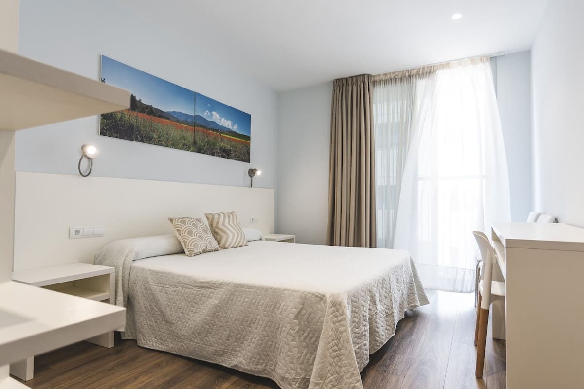 Habitación de hotel moderna con cama, escritorio y una silla. Se observa un cuadro en la pared y una ventana con cortinas.
