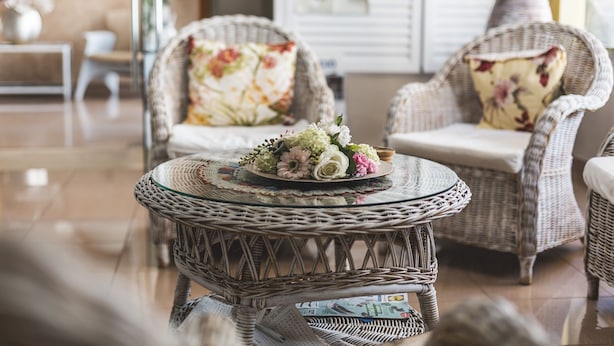 A wicker chair with a floral cushion, a glass table with a floral arrangement, and a magazine on the floor.