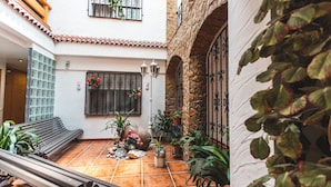 A courtyard with a tiled floor, a bench, potted plants, a window with bars, and a stone wall.