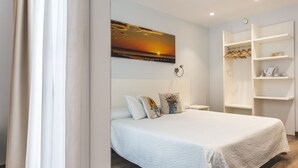 A modern bedroom with a large bed, a nightstand, a wall-mounted lamp, and a painting of a sunset.