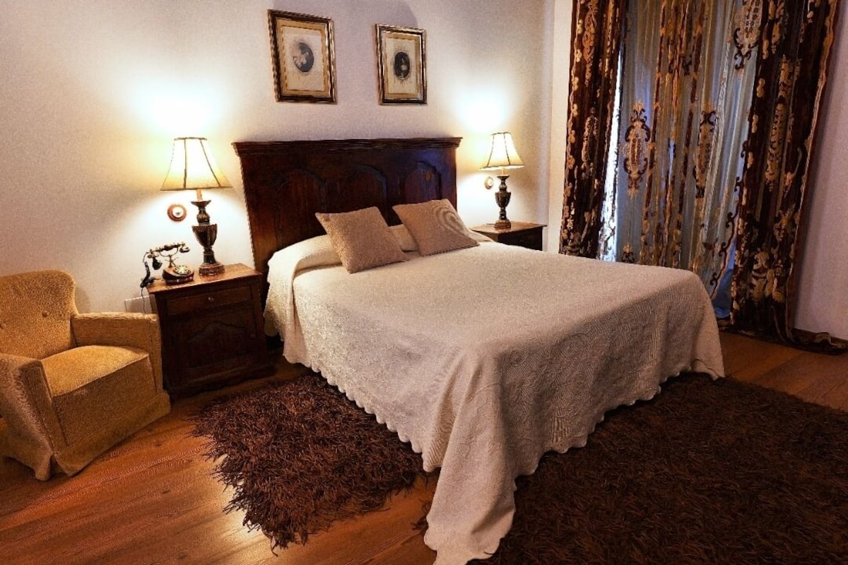 Una cama bien hecha con una colcha blanca y dos almohadas, flanqueada por dos mesitas de noche con lámparas. Un sillón marrón está situado a la izquierda de la cama.