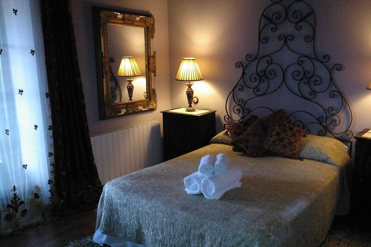 Una cama bien hecha con una toalla blanca enrollada, un cabecero decorativo, dos lámparas de noche y un espejo en la pared.