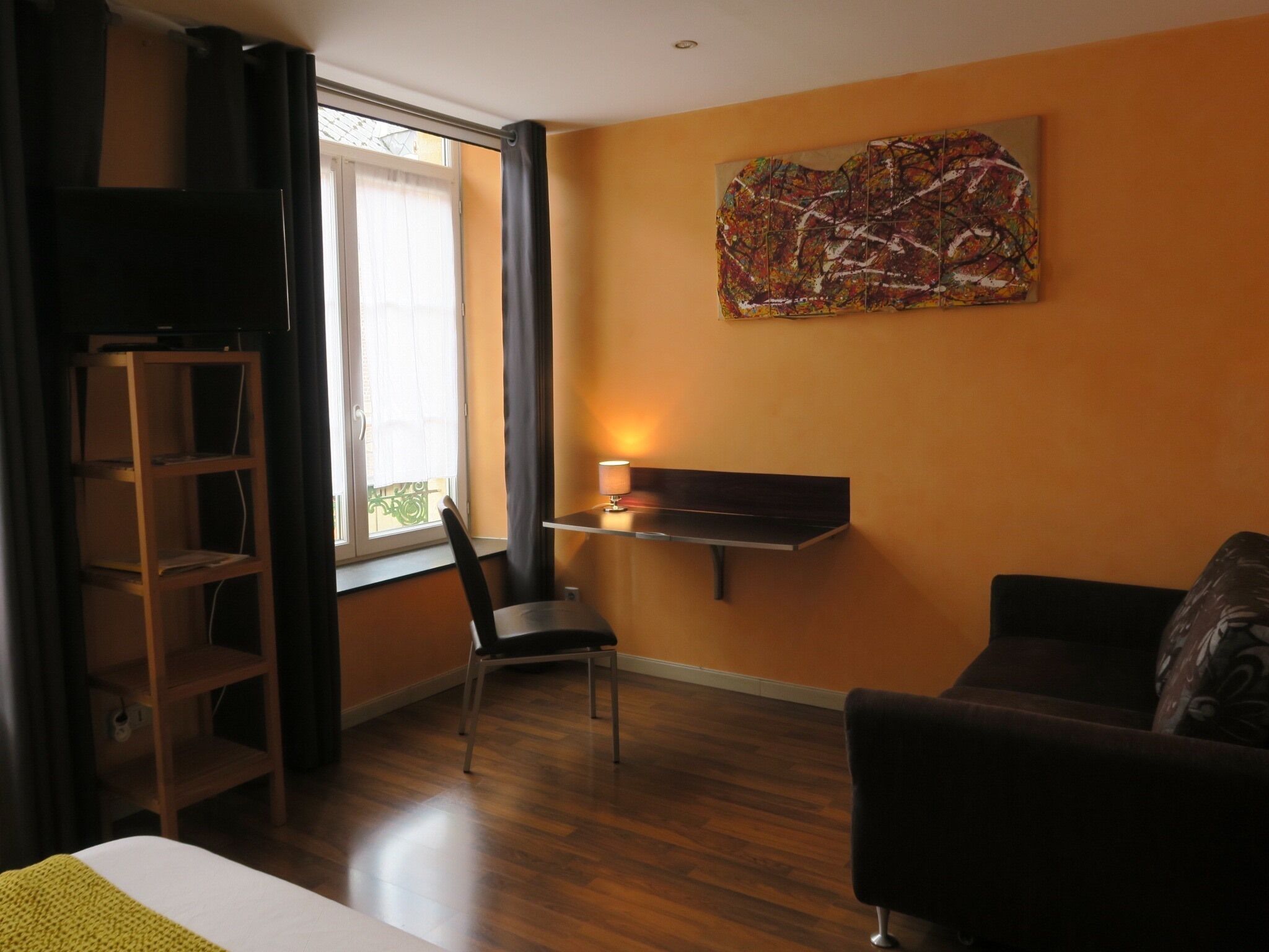 Une pièce avec un plancher en bois, un bureau avec une lampe, une chaise, un canapé, un téléviseur posé sur un meuble et un grand tableau abstrait accroché au mur.