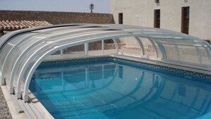 A swimming pool with a glass enclosure, surrounded by a stone wall and a building with windows. - Casa Rurales Ventaseca (Mazarron)