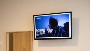 Een kleine kamer met een houten bureau, een aan de muur bevestigde televisie, een witte waterkoker en een houten kast.