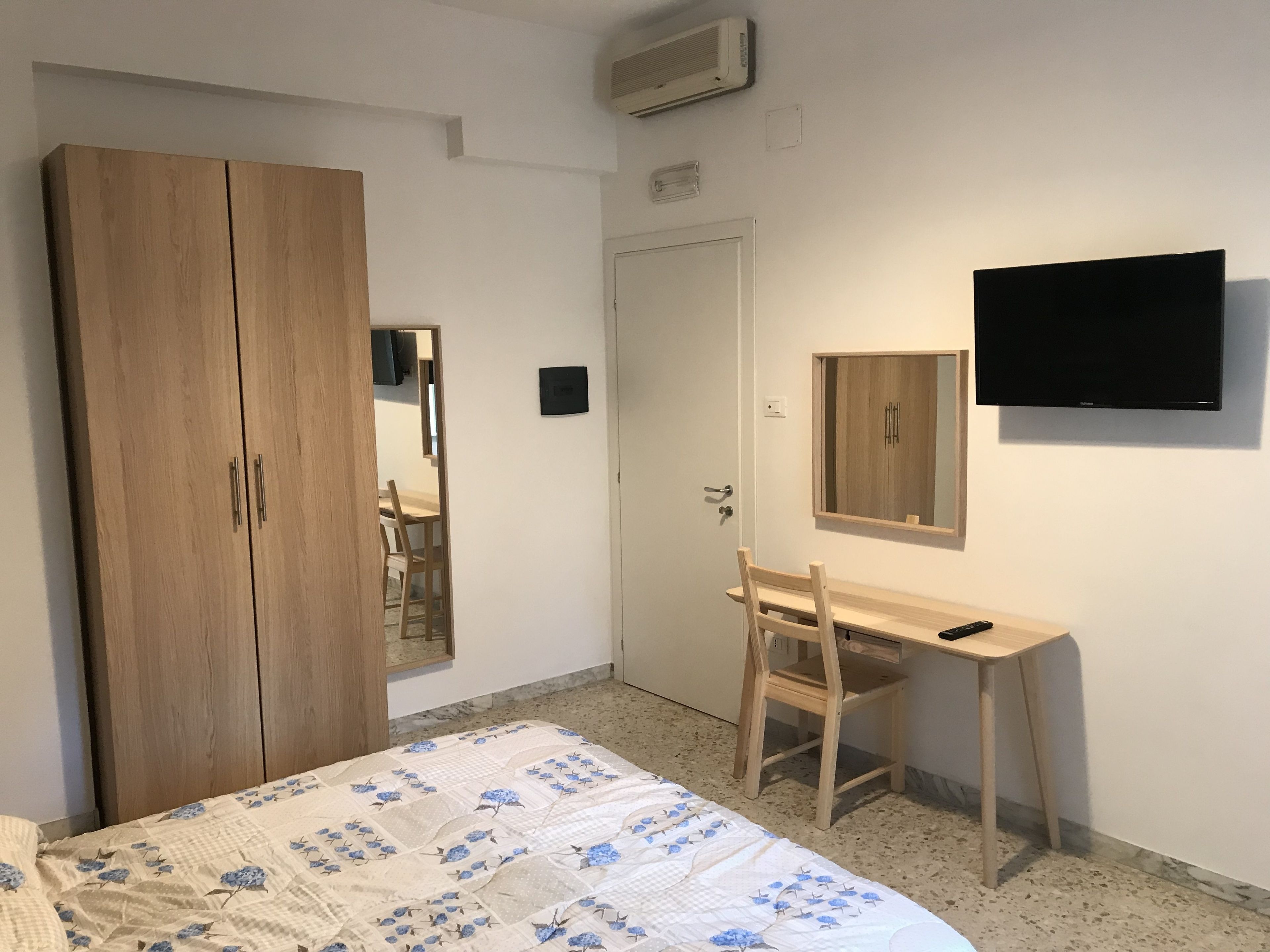 A bedroom with a bed, a wooden wardrobe, a desk with a chair, a television, and a mirror.