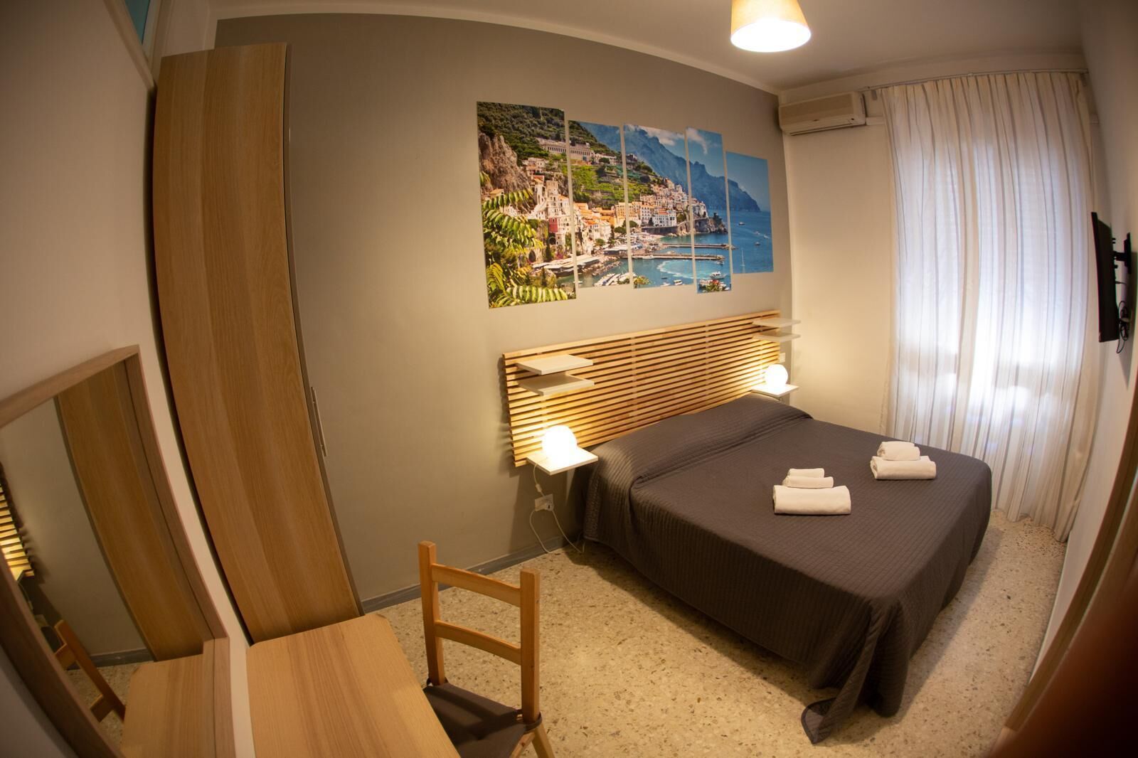 A hotel room with a bed, bedside table, chair, and a large painting of a coastal town.